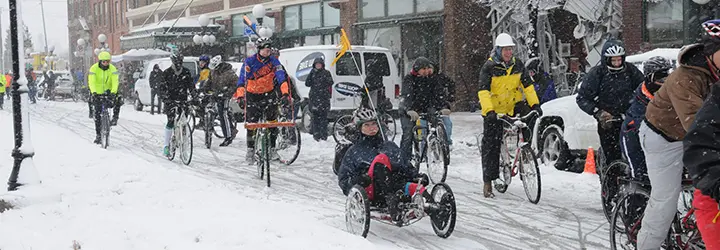 A group of bikers pedal through a snow-covered main street. 