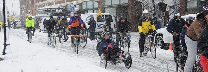 A group of bikers pedal through a snow-covered main street.