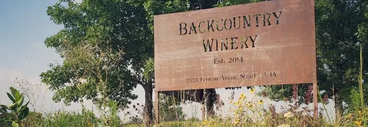 A sign that says, "Backcountry Winery" stands among green trees and grass.