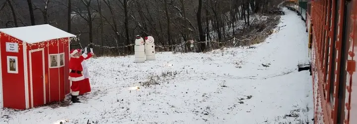 Santa and Mrs. Claus stand before a red hut waving to a passing train.