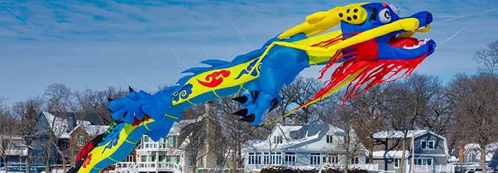 A bright blue, red and yellow oriental dragon kite flies above a frozen lake and the crowd below.