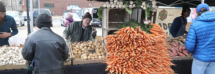 A produce vendor gives a thumbs up behind his stand filled with carrots, yams and onions. 