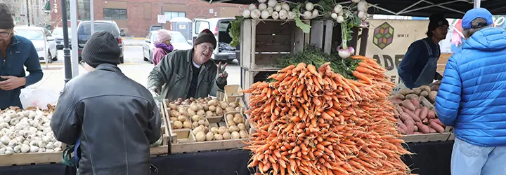 A produce vendor gives a thumbs up behind his stand filled with carrots, yams and onions.