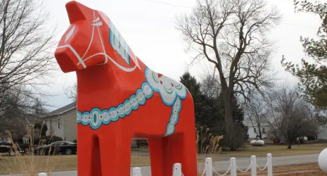 A bright red Dala Horse stands in front of bare trees and a gray sky.