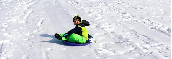 A boy wearing bright green/yellow snowpants and coat races down a hill on a circular sled.