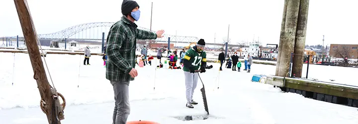 Two men work to cut a piece of ice from a frozen river as a group of specators watches from behind them. 