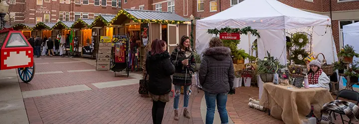 Bundled up shoppers enjoy a cup of coffee in front of holiday craft huts.