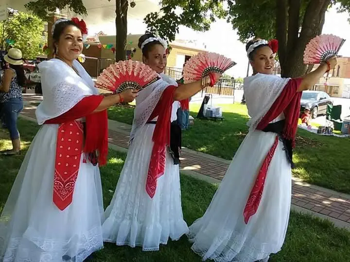 Three Latina dancers in white dresses hold up red oriental fans and pose for the camera. 