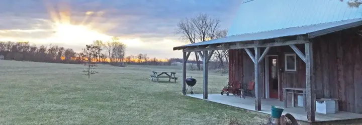 A small, faded red barn with a cement covered porch as the sun sets behind it.