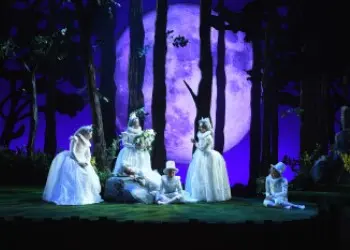 Opera singers donning white dresses and suits sing before a forested backdrop illuminated by a full moon.