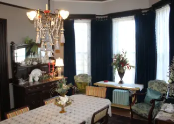 A historic dining room with a cloth-covered table, a dark wood hutch, a chandelier with tassels and bay windows.