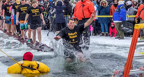 A man wearing shorts and a t-shirt jumps into a hole cut into a frozen lake as safety personnel and a crowd watch.