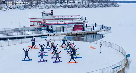 A yoga group stretches on a frozen pier before a boat floating on a frozen lake.
