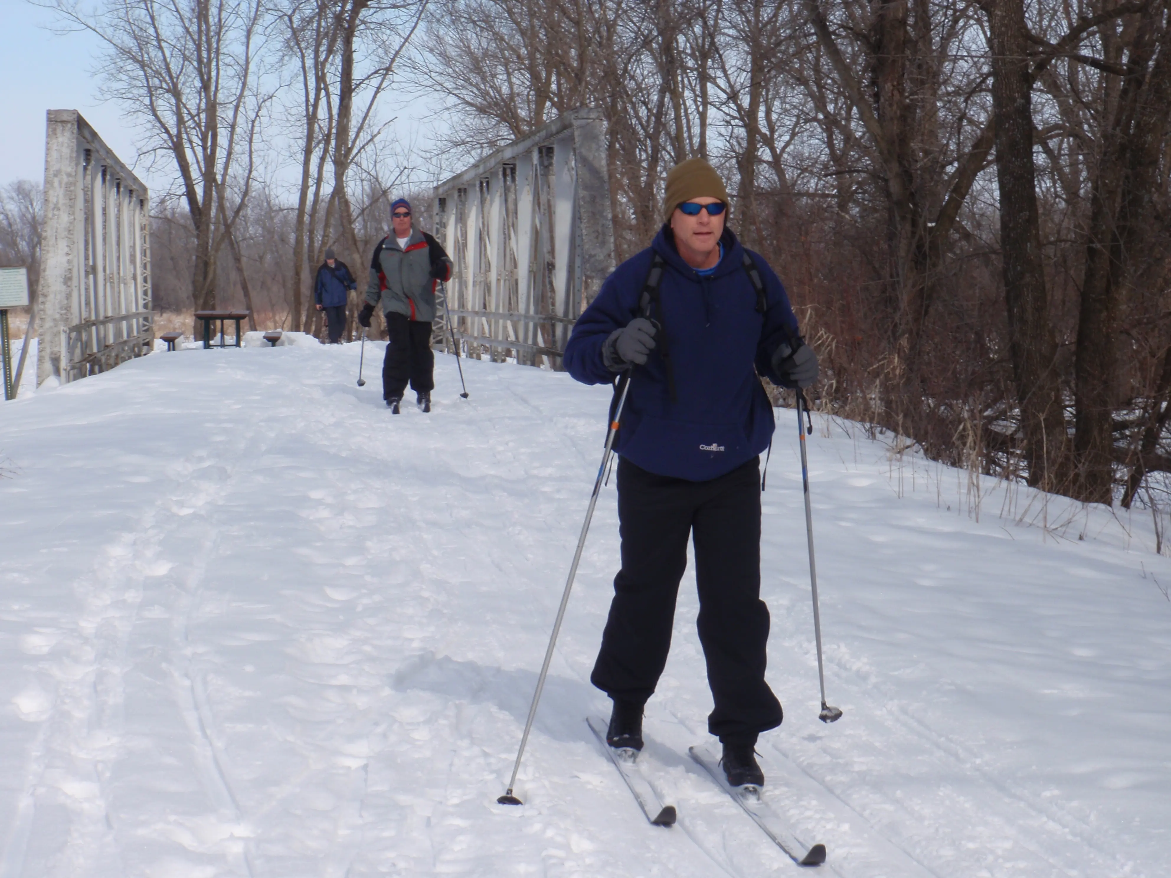 An older white man cross-country skis as other skiers cross a bridge behind him.