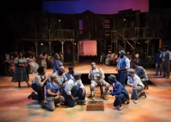 A group of Black opera performers dressed in 70s clothing gather around a member.