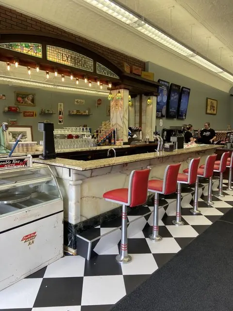 An old soda fountain with a marble counter, red stools and a black and white tiled floor.