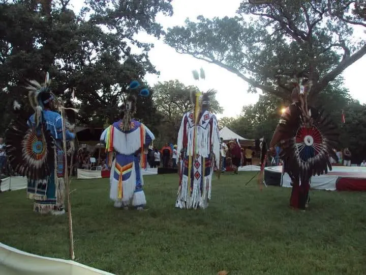 Four Native American dancers stand before a crowd in their traditional clothing.