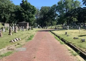 An old red brick road leads through a cemetery filled with old headstones and green trees.