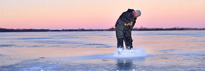 A man cuts through the ice to prepare for ice fishing as a pink sunset floods the sky behind him.