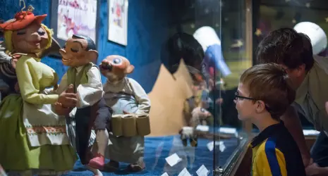 A boy wearing glasses gazes at an intricate marionette couple embraced in dance.