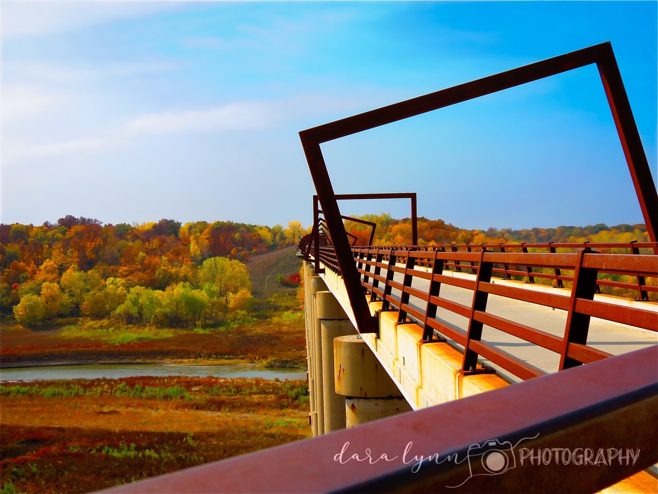 A steel bridge stretches across a vibrant river valley filled with fall foliage.