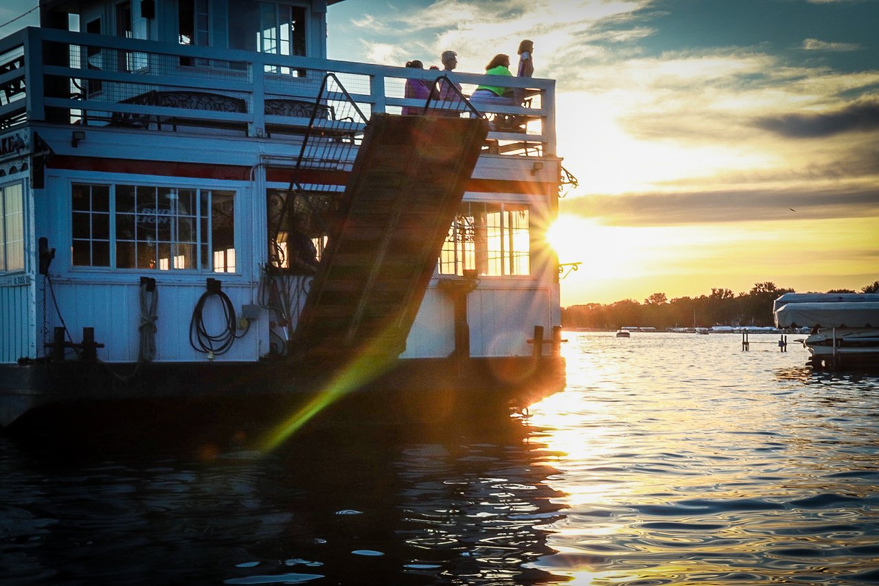 A historic riverboat sits in the water as the sun sets behind it.