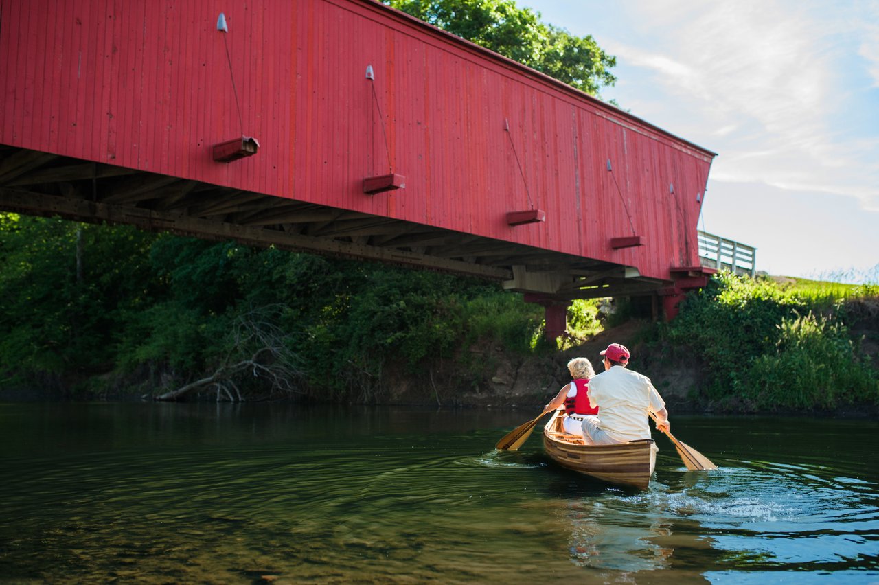 A father and daughter canoe beneath a red covered bridge.