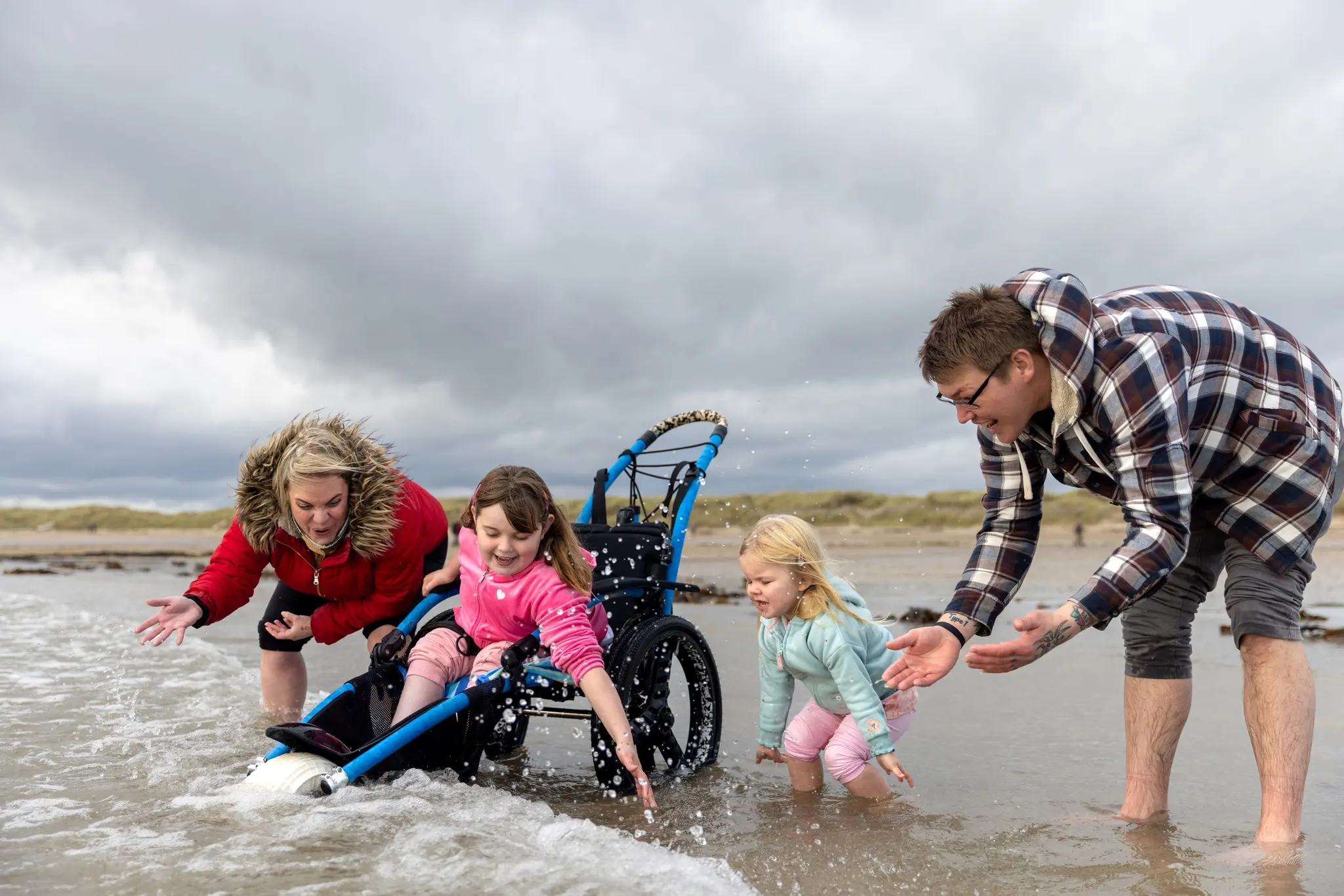 A young girl in a wheelchair splashes in the water at a beach with her mom, dad and younger sister.